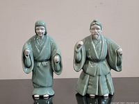 Pair of celadon-glazed Japanese porcelain figurines, standing side by side on a reflective surface. Both approximately 7.5 inches tall.