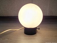 White glass globe lamp illuminated on a black metal base showing its shape and size.