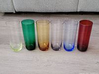 Front view of all six highball glasses in varied colors arranged in a row on light wood flooring in front of a couch.
