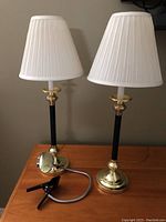 Two bedside lamps with black and gold bases, cream pleated lampshades, placed on wooden surface.