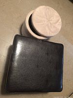 Photo showing top view of round beige/cream faux suede ottoman and top of square dark brown faux leather ottoman.