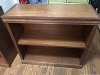 Photo showing one of two walnut stained wooden bookcases with two shelves each, placed on wood flooring, highlighting the top molding and grain.