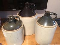Photo showing three old crockery jugs of different sizes on a wooden surface, with TV screen and other household items in background, illustrating vintage ceramic jug styles with glazed tops and stoneware bases.