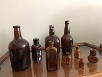 Photo showing nine brown antique glass bottles of various shapes and sizes arranged on a wooden surface.