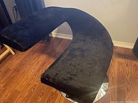 Black curved chair with black faux velvet upholstery and shiny chrome base on wood flooring.