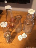Four clear glass dairy milk bottles of various shapes on a wooden surface, one with a red Acme Dairy print, one containing wooden toothpicks, others plain or wide-mouthed, all topped with various white plastic caps.