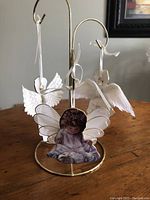Three porcelain angel ornaments hanging from a gold metal stand, showing front views with detailed angel illustrations and white ribbon loops.