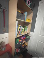 Photo of a tall light wood finish IKEA bookcase with four shelves containing various children's books and plastic storage bins.