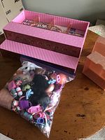 Clear resealable plastic bag holding numerous small LOL Surprise doll accessories such as shoes and bottles on a wooden table beside the pink display case.