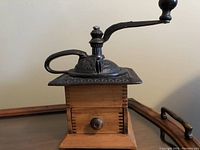 Antique wooden pepper mill featuring a wooden body with dovetail joints, metal crank handle, and decorative metal lid.