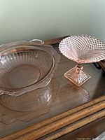 Pink depression glass bowl or plate with handles and ribbed floral pattern, placed on wooden surface.