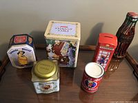 Collector tins of various classic brands arranged on table: Uneeda Biscuit, Hershey's Milky Mix, Hershey's Cocoa, Uncle Joe's Mint Balls, Harrods telephone booth, and Coca-Cola bottle-shaped coin bank.