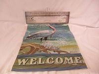 Full view of double sided garden flag with heron design and 'WELCOME' text, partially extending from packaging.