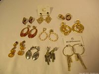 Photo showing all 10 pairs of earrings laid out on a white surface, featuring a variety of styles including gold-tone hoops, red enamel drops, and silver-tone dangles.