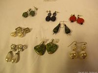 Eight pairs of assorted vintage earrings arranged on white surface including green stone hearts, faux pearls, and colorful drops.