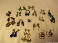 Photo showing all earrings in the lot arranged on a white fabric background. Various styles and colors visible including green Christmas tree shapes, blue teardrop shapes, gold and crystal fringes, and black bead clusters.