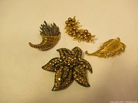 Four vintage gold-tone brooches laid out on a flat surface including cornucopia, berry cluster, leaf, and flower shapes.