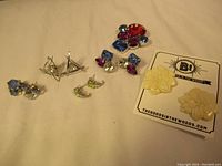 Photo of six pairs of earrings arranged on a white surface showing variety of styles and colors, including vintage cream floral clip-ons on a branded card and multi-colored rhinestones.