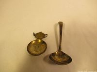Two antique spoons, one teapot-shaped handle with floral engraving, one ladle style spoon, both aged metal with patina.
