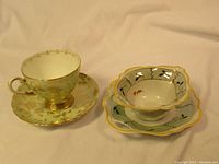 Photo showing two vintage Coalport teacups on their matching saucers. One set is gold and green floral, the other is a scalloped white cup with black and red floral design and yellow rim.