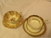 Two vintage Paragon teacups on matching saucers, showing floral design and gold filigree on one set and gold geometric pattern on the other.