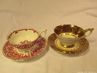 Two vintage china teacups with matching saucers, one Royal Stafford red floral design and one Coalport burgundy and gold ornate design, side by side.