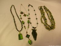 Four necklaces laid out on a beige background, showing various green pendants and beads including a large rectangular pendant, earrings, a dark green arrowhead pendant, and chip bead necklace.