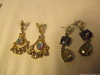 Pair of gold-tone earrings with iridescent teardrop and multiple dangling rhinestones, and silver-tone earrings with dark crystal tops and abalone shell drop dangles on a beige background.
