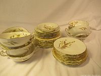 Full view of set showing multi-handled bowls, stack of plates and smaller bowls with gold wheat and leaf decoration and gold trim on edges.