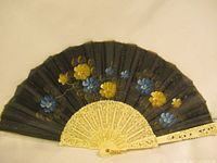 Front view of the vintage folding hand fan showing the hand-painted floral design in blue, yellow, and gold on black fabric, with cream-colored intricately carved sticks and guard.