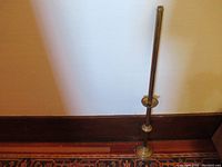 Full view of tall vintage brass church candlestick resting on the floor against a wall, showing the round tiered base and slender shaft with two circular platforms.