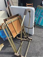 Photo of multiple folding tables with white plastic tops and gold metal legs, stacked and leaning against one another with other chairs in background.