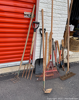 Photo shows a variety of garden tools including rakes, a hoe, shovels, and a pick axe all with wooden handles leaning against a wall.
