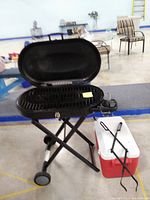 Open charcoal grill on folding wheeled stand next to red and white cooler and black log tongs