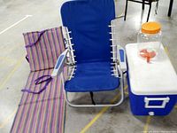 One photo of the lot showing a blue padded beach chair with armrests, a striped foldable beach mat or chair, a clear plastic water jug with orange lid and spigot, and a blue Coleman cooler with white lid and handles, all placed on a floor with marking tape.