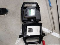 Fish finder in black carrying case with instruction manual included, shown on concrete floor with shoe partially visible.