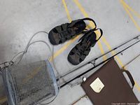 Men's water shoes, cylindrical minnow cage, brown seat, fishing rods and reels on concrete floor
