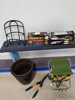 Photo showing black metal hose caddy, two boxed Moonrays solar lights, dark brown plastic planter, gardening shears with green and yellow handles, and green padded gardening stool with foldable legs and storage pouch.