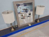 Pair of table lamps flanking a rectangular wall mirror against a blue wall and grey flooring.