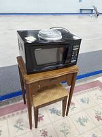 Black Sylvania microwave on wooden sewing machine table with stool underneath, placed on pink patterned rug