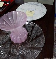Pink shell-shaped glass vase, large glass platter with swirl texture, and marble lazy susan on a wooden surface
