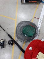 Photo showing black fishing rod with black foam handle and spinning reel, beside green wire mesh fish cage with green plastic cover and metal frame, along with red thermal carry bag.