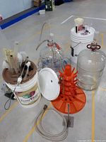 Photo showing two glass carboys with airlocks and red valves, white and brown fermentation buckets, clear tubing, and an orange drying rack.