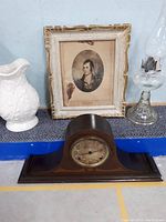 Full lot image showing white ceramic water jug, framed portrait, mantle clock, and glass oil lamp arranged on blue shelf.