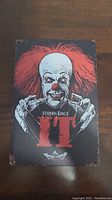 Metal sign featuring Pennywise the Clown from Stephen King's It with red hair and clown makeup, distressed edges and background.