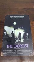Photo shows an 8x12 inch metal sign featuring The Exorcist movie poster artwork with a dark scene of a man standing in front of a house under a streetlamp.