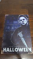 Rectangular metal sign with Michael Myers holding a knife in front of a house, text 'John Carpenter's HALLOWEEN' visible