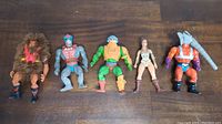 Five MOTU He-Man figures arranged on wooden floor showing full bodies.