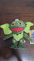Full front view of the green Stripe Gremlin plush toy with wing-like ears, yellow-orange eyes, and red-toothed mouth, displayed on a wooden surface.