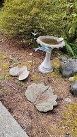 Outdoor setting showing concrete birdbath filled with water, two leaf decorations on ground (one broken), metal butterfly and fish yard art in natural garden environment.